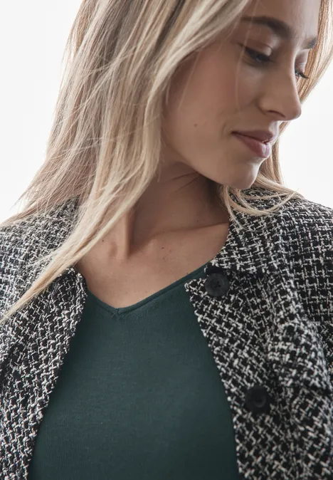 Frau mit blondem Haar trägt grünes Shirt und karierte Jacke, schaut leicht nach unten.
